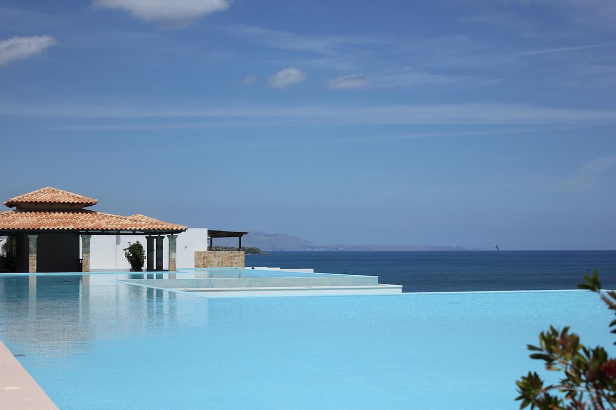 A modern white-walled boutique hotel pool surrounded by palm trees and wooden loungers reflects a soft late afternoon sky