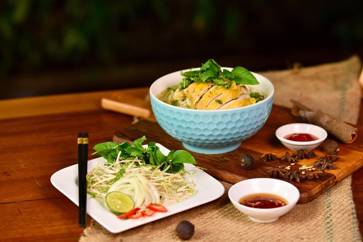 A steaming bowl of bun mam with rice noodles, shrimp, slices of pork, and tropical herbs sits on a plastic stool in a Can Tho market